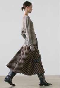 Langärmliges, hellgraues Oberteil mit Twist-Detail, kombiniert mit einem braunen karierten Maxirock. Das Modell trägt kniehohe schwarze Stiefel und hält eine kleine Tasche.