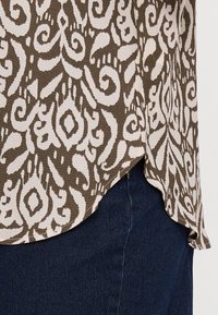 Top marrone con motivo astratto bianco, con orlo arrotondato e tessuto testurizzato, abbinato a jeans scuri.