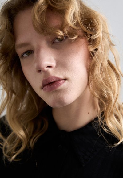 Prim-plan al unei persoane cu păr blond ondulat de lungime medie. Aceasta poartă o cămașă neagră cu guler, cu un model texturat, având nuanțe naturale ale pielii.