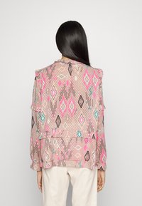 Une femme avec de longs cheveux noirs porte un blouse rose clair ornée de motifs géométriques colorés et de détails volants, associée à un pantalon crème, de dos.