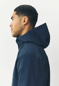 Chaqueta con capucha en color azul marino, de cuello alto, textura suave y diseño minimalista. Presenta un corte relajado y detalles de costura sutiles.
