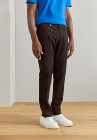 Pantaloni neri a taglio slim, abbinati a una camicia blu e scarpe da ginnastica bianche, in piedi su un pavimento in legno contro uno sfondo neutro.
