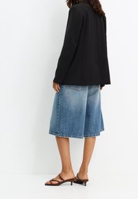 Blazer noir sur mesure, culottes en denim bleu ample avec bords délavés, et sandales marron à lanières. Tissu lisse et silhouette décontractée.