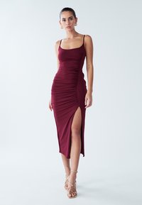 Vynuogių spalvos kūno formą pabrėžianti suknelė su raukiniais, vidurinio šlaunies kirpimo plyšiu ir plonais spaghetti petnešėlėmis; lygi, elastinga medžiaga.