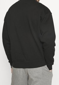 Svart sweatshirt med strukturerat tyg, rund halsringning och ribbade muddar; bärs med gråa mjukisbyxor och visar en avslappnad passform.