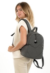 Junge Frau trägt eine ärmellose weiße Fleeceweste und beige Hosen, mit einem dunkelgrauen, gesteppte Rucksack über der Schulter.