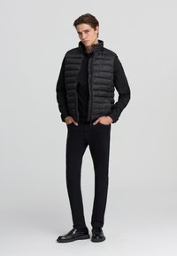 Czarna kurtka puchowa z zamkiem z przodu i wysokim kołnierzem, noszona na czarnej koszulce z długim rękawem. W komplecie czarne spodnie slim-fit i buty.