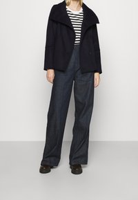 Chaqueta de lana azul marino con un cuello ancho y dos botones, combinada con una camisa a rayas y pantalones de mezclilla oscuros holgados. Zapatos negros.