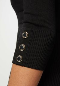Manche de pull en tricot noir avec trois boutons dorés décoratifs le long d'un poignet côtelé, présentant une texture lisse et un design ajusté.