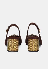 Zapatos slingback de ante marrón con un tacón decorativo adornado con tachuelas doradas. El tacón presenta un patrón de cuadrícula de brillantes embellishments dorados.
