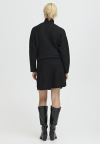 Zwarte gebreide trui met een hoge kraag, gecombineerd met een bijpassende zwarte minirok. Glanzende kniehoge zwarte laarzen maken de outfit af.