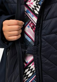 Giacca trapuntata blu navy con zip, che rivela uno strato interno decorato con disegni geometrici in rosa, blu e nero su uno sfondo bianco.