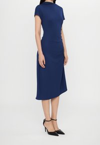 Mörkblå figursydd klänning med korta ärmar, draperad detalj i sidan och asymmetrisk fåll, kombinerad med svarta spetsiga högklackade skor.