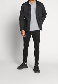 Chaqueta negra con dos bolsillos frontales, camiseta gris, jeans skinny negros y zapatillas deportivas negras. El material parece de tela, texturizado, diseño casual.