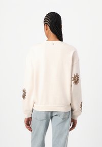 Beige sweatshirt with long sleeves, featuring brown embroidered sun designs on the sleeves and a simple round neckline.