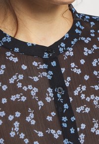Durchsichtiges schwarzes Oberteil mit floralem Muster in Hellblau, runder Ausschnitt und zwei schwarzen Knöpfen auf der Vorderseite.
