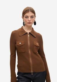 Cárdigan marrón de canalé con cremallera y cuello, dos bolsillos frontales y acentos de botones dorados. Textura suave y diseño ajustado.