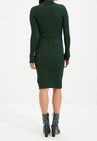 Abito verde a maglia a coste con collo alto e maniche lunghe, silhouette aderente, che arriva a metà polpaccio. Abbinato a stivali alla caviglia verde scuro.