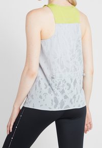 Canotta sportiva grigio chiaro con una texture liscia, presenta una parte superiore verde lime e un motivo perforato su tutta la superficie, abbinata a leggings neri.