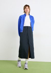 Blaue Zip-Jacke, weißes T-Shirt, schwarzer knöchellanger Rock mit seitlichem Schlitz, weiße Socken und graue Sneaker. Grüner Boden, schlichter Hintergrund.