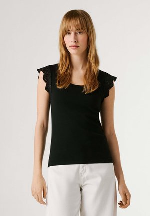 Jeune femme aux longs cheveux châtain clair portant un haut noir sans manches avec des détails en dentelle et un pantalon blanc, debout devant un fond uni.