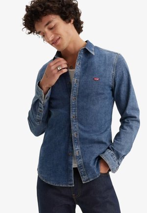 Jeune homme aux cheveux bouclés portant une chemise en jean bleu Levi's, un haut blanc en dessous, un pantalon foncé et une bague au doigt.