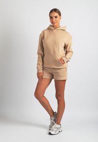 Beige-huppari, jossa on etutasku, yhdistetty samanvärisiin shortseihin. Pehmeä materiaali ja rento istuvuus. Yhdistetty valkoisiin ja harmaisiin urheilukenkiin.