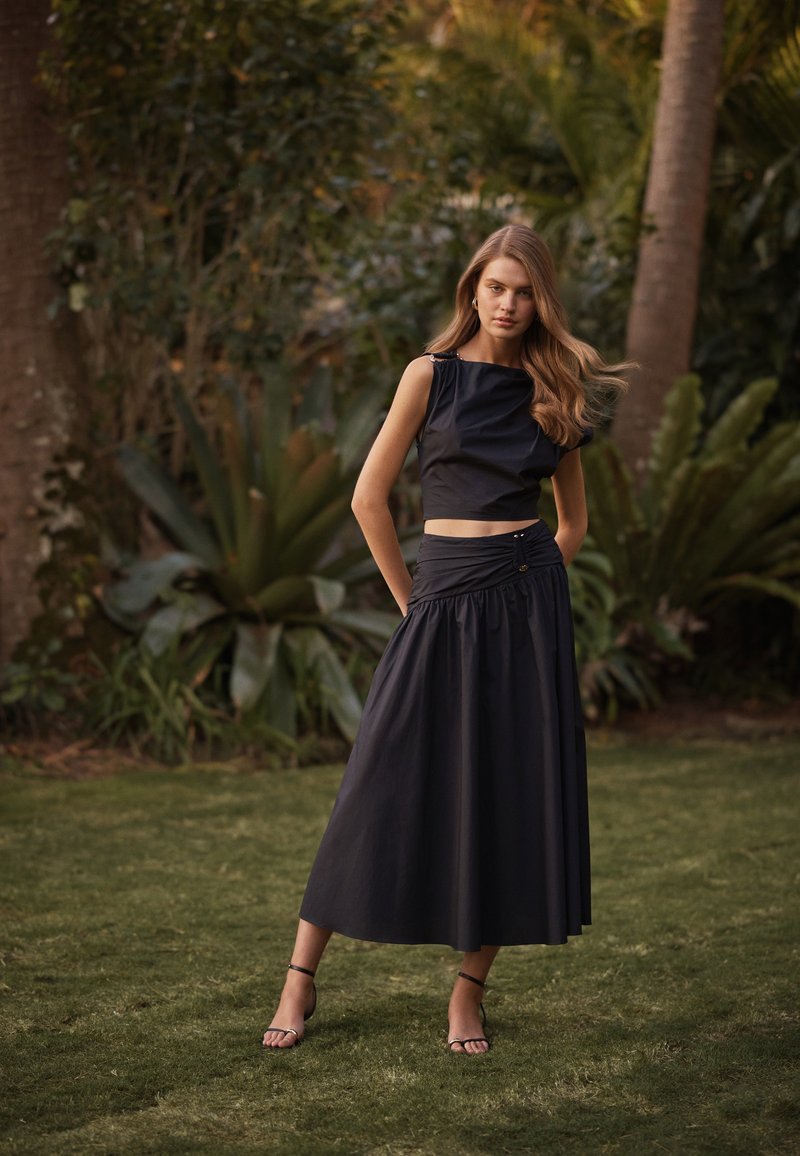Γυναίκα που φοράει ένα μαύρο αμάνικο crop top και ταιριαστό midi φούστα με ψηλή μέση, στέκεται στο γρασίδι με φυλλώδη φυτά στο φόντο.
