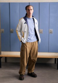 Chaqueta blanca con cremallera y rayas amarillas, camiseta de rayas azules y pantalones cargo beige. Zapatos marrón oscuro. Taquillas azules en el fondo.