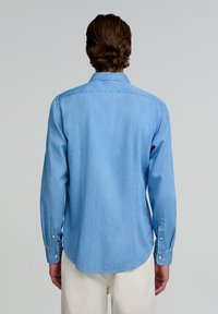 Cămașă din denim bleu deschis, cu mâneci lungi, guler ascuțit și manșete cu nasturi. Textură netedă și detaliu cu tiv curbat la partea de jos.
