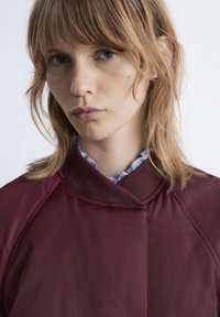 Bordeauxfarbene Bomberjacke mit hohem Kragen, kontrastierender Stoffstruktur und glatter Oberfläche. Darunter ist ein hellblaues Hemd getragen.