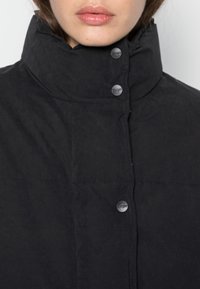 Primer plano de una persona vestida con una chaqueta negra de cuello alto, con botones a presión negros visibles y detalles de costura mínimos.