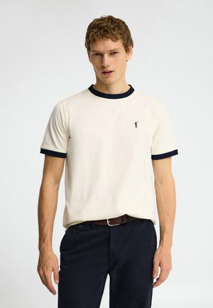 Hombre con camiseta blanca de manga corta con ribete negro en el cuello y las mangas, pantalones negros y cinturón marrón, de pie sobre un fondo liso.