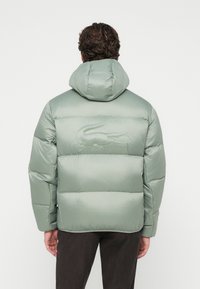 Jachetă puffer verde deschis cu glugă, având un design cusut și un subtil logo de crocodil embosat pe spate.