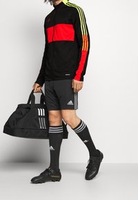 Chaqueta deportiva negra con rayas rojas y amarillas, pantalones cortos grises, calcetas negras con rayas blancas y botas de fútbol negras. Lleva una bolsa negra.