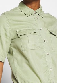 Camisa de manga corta verde claro con cuello, dos bolsillos en el pecho y botones a presión plateados. Tejido suave y liso con una ligera textura brillante.