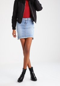 Denim-Bleistiftrock mit ausgefranstem Saum, kombiniert mit einer schwarzen Bomberjacke und einem roten Oberteil. Schwarze, glänzende Ankle Boots mit strukturiertem Design.