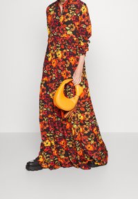 Dlouhá černá květinová šaty s červenými, oranžovými a žlutými květy; má límec, dlouhé rukávy a nařasený pas. Drží malou oranžovou tašku.