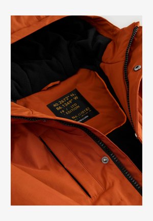 Orange isolierte Jacke mit schwarzem Fleece-Futter, ausgestattet mit einem Frontreißverschluss, Druckknöpfen und einem Innenetikett mit Koordinaten und Editionsnummer.