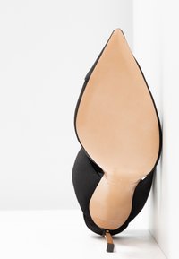 Chaussure à talon haut noire à bout pointu reposant sur son talon, montrant une semelle marron claire lisse et un talon aiguilles fin sur fond blanc.
