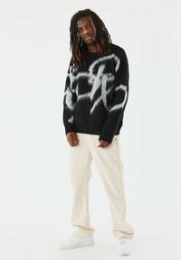 Pull noir flou avec un motif abstrait blanc, assorti à un pantalon couleur crème. Des baskets blanches viennent compléter le look. Les contrastes de texture sont évidents.