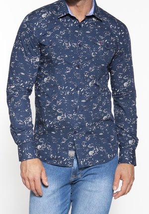 Camisa de botones azul marino con patrones abstractos en blanco, con cuello, mangas largas y detalle de bolsillo frontal. La tela parece suave.