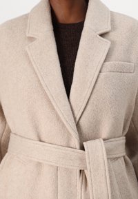 Cappotto di lana beige con finitura testurizzata, rever a punta, un'unica tasca sul petto e una cintura in tessuto legata in vita.