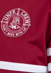 Červený dres s kulatým logem, které obsahuje nápisy "UNFR CREW" a "DON'T MESS WITH US" v bílém vyšívání, doplněný bílými pruhy.