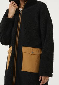 Person med svart strukturerad fleecetröja med frontdragkedja och två stora tanfärgade ficklock med tryckknappar.