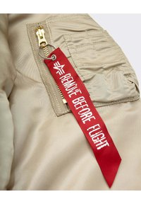 Beige nylon fabric with a gold zipper and a red “REMOVE BEFORE FLIGHT” tag featuring white text and a logo. Smooth texture, functional design.