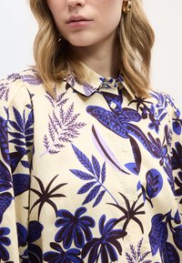 Γυναίκα που φοράει κρεμ πουκάμισο με κουμπιά, με μπλε και σκούρα καφέ τροπικά φύλλα και χρυσούς κρίκους στα αυτιά.