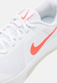 Zapatilla deportiva blanca con parte superior de malla, superficie texturizada, acento en coral en el lateral y cordones blancos. Cuenta con una suela de goma para mayor tracción.