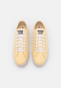 Un paio di sneakers low-top gialle Converse All Star con lacci bianchi e suole in gomma, viste dall'alto su uno sfondo bianco.