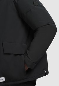 Crna izolirana jakna s rukom u bočnoj đepu, vidljive oznake marke na rubu i bočnom šavu, te okrugla oznaka logotipa na gornjem rukavu.